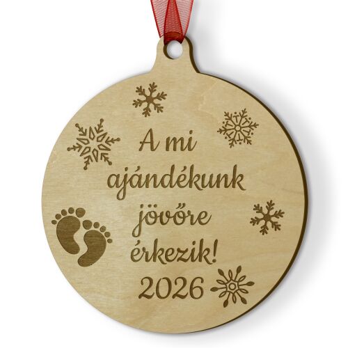 Közelkép egy fából készült karácsonyfadíszről, amely egy baba 2026-os érkezését hirdeti, hópelyhekkel és baba lábnyomokkal gravírozva