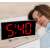 Digital alarm clock with curved mirror display and red LED, showing a woman sleeping in the background