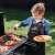 Child wearing a black grill apron with red trim, grilling food on a barbecue