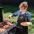 Child wearing a black grill apron with red trim, grilling food on a barbecue