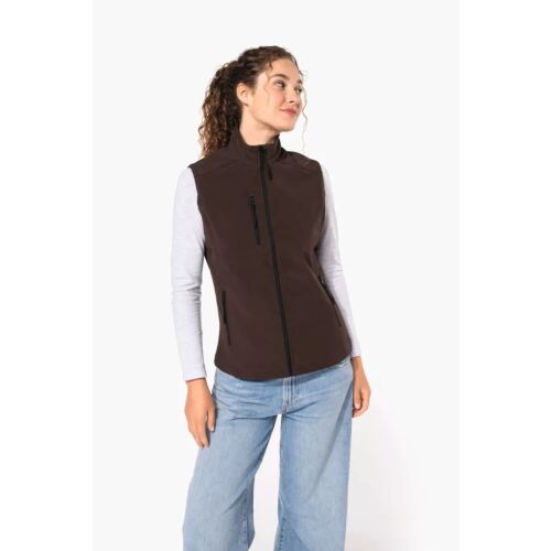 Nő visel barna Kariban KA404 softshell mellényt fehér hosszú ujjú felsővel és farmerrel, alkalmas hétköznapi és kültéri viseletre - Női mellény
