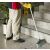 Takarító söpör egy szürke pettyes padlót egy Vileda Professional Hard Eco 30 cm-es seprűvel egy beltéri kereskedelmi környezetben, lépcső és növény közelében, bemutatva a használatát
