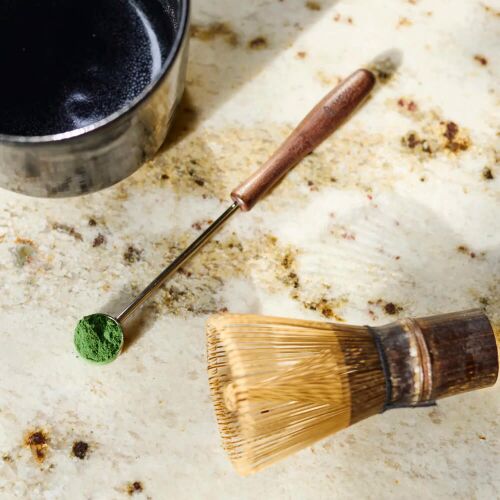 Moya Matcha rozsdamentes acél adagolókanál zöld matcha porral és habverővel egy pöttyös munkalapon, ideális a pontos matcha elkészítéséhez. - Konyha és étkezés