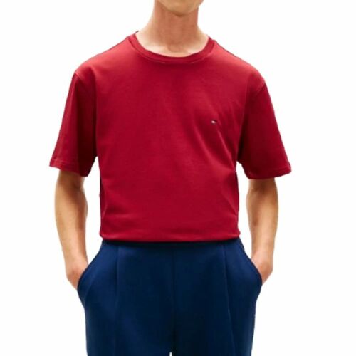 Elölnézet egy férfiról, aki piros Tommy Hilfiger FLAG TEE pólót visel kis logóval, kék nadrággal, fehér háttér előtt, bemutatva klasszikus szabását. 