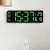 Digital wall clock with green LED display showing time, date, day of the week, and temperature, mounted on a wall